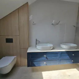 Nowoczesna łazienka z szafką pod umywalki w kolorze granatowym i drewnianą zabudową pod skosem. Umywalki owalne, białe. Na ścianie płytki ułożone w jodełkę.