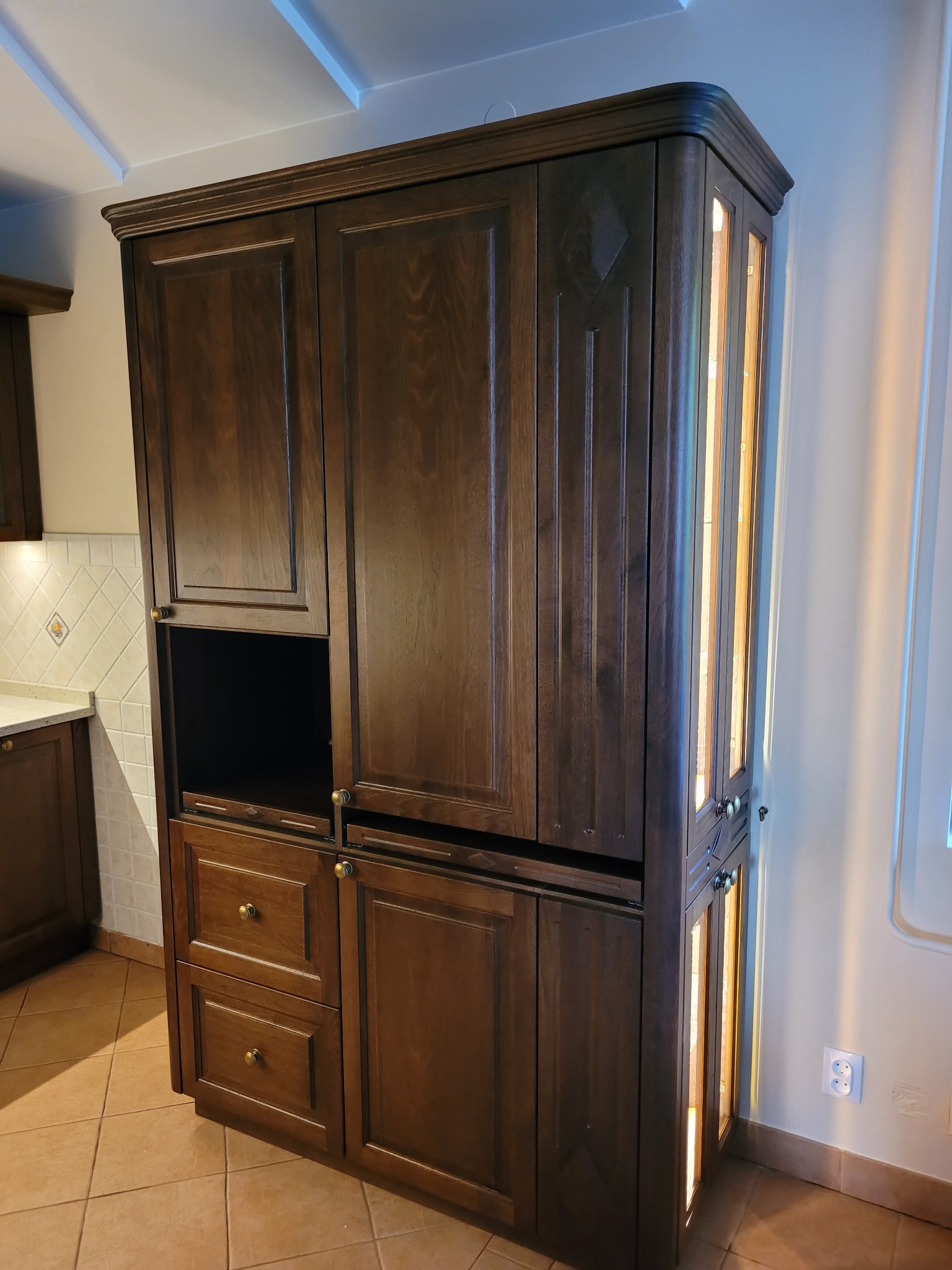 Masywna, drewniana szafa w zabudowie kuchennej z oświetleniem wewnętrznym, dwiema szufladami i otwartą półką na sprzęt kuchenny, widoczna płytka ceramiczna na ścianie i gniazdko elektryczne.