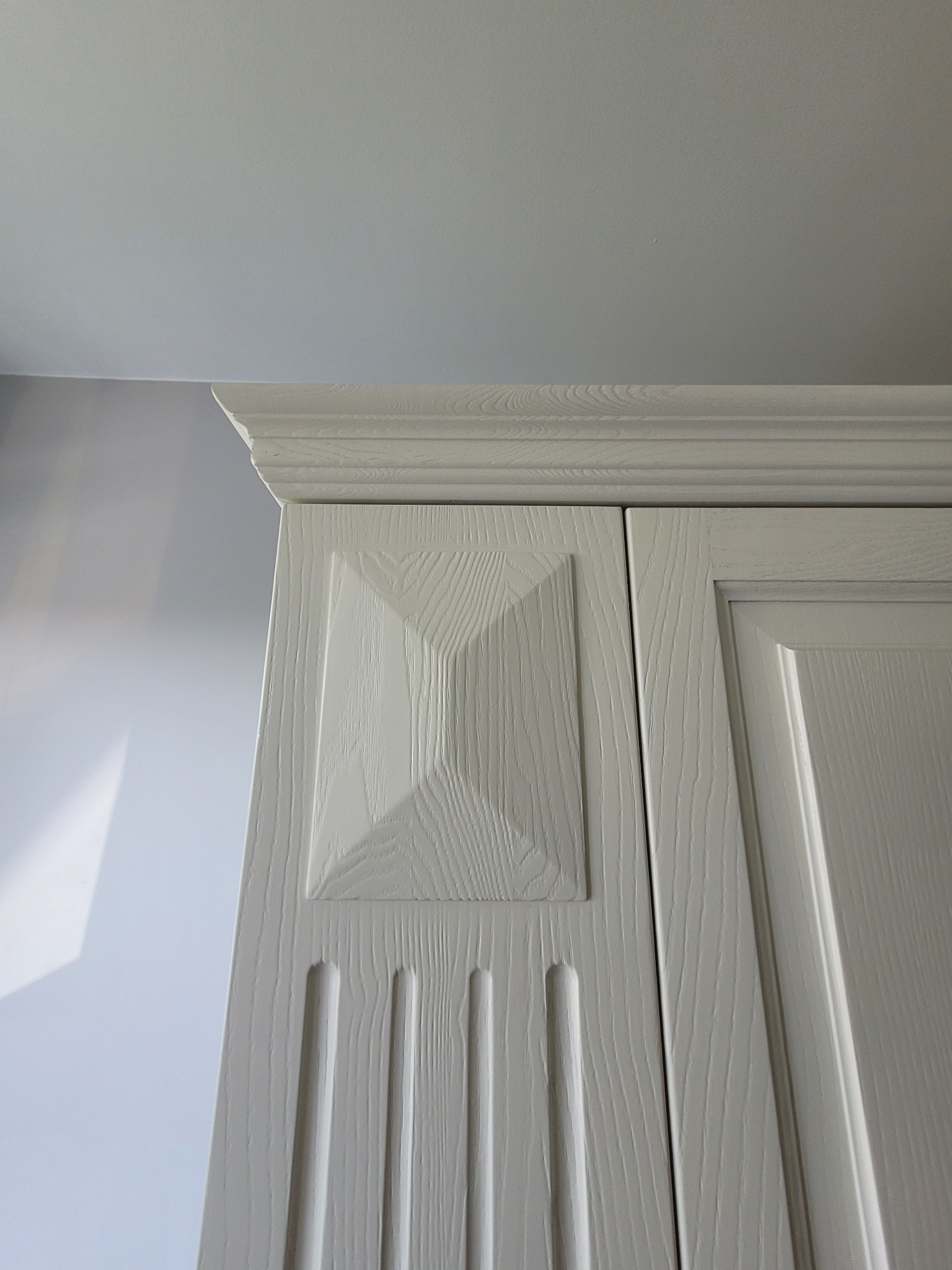 Fragment białej szafy z frezowanymi drzwiami i ozdobnym zwieńczeniem, widoczna struktura drewna.