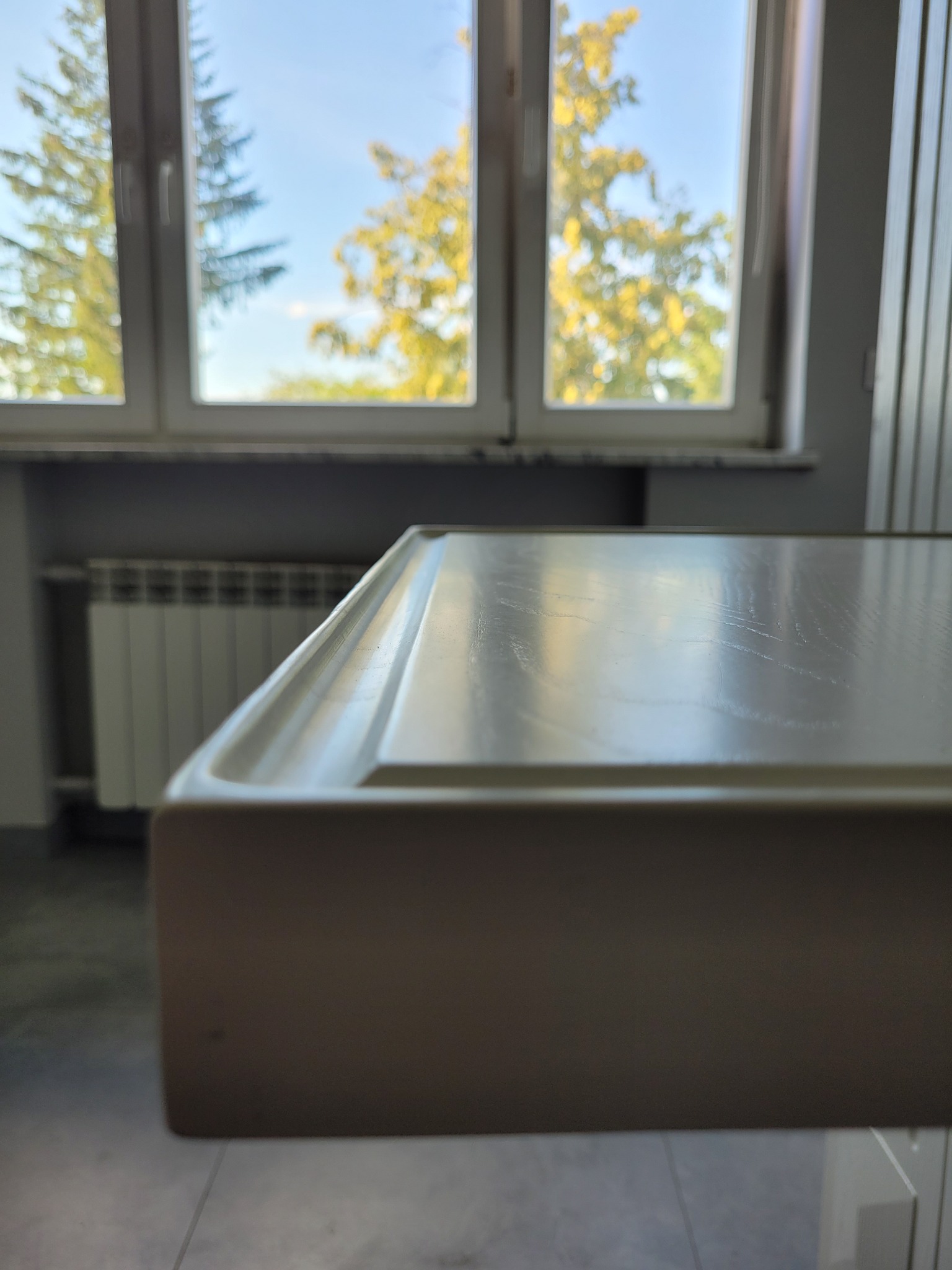 Fragment blatu w jasnym odcieniu z widoczną strukturą drewna, w tle okno z widokiem na zieleń i grzejnik.