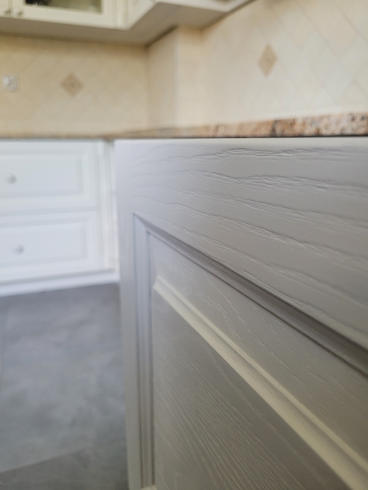 Szary front szafki kuchennej z widoczną strukturą drewna, w tle beżowe płytki i białe szafki, ujęcie z bliska podkreślające detale wykończenia.