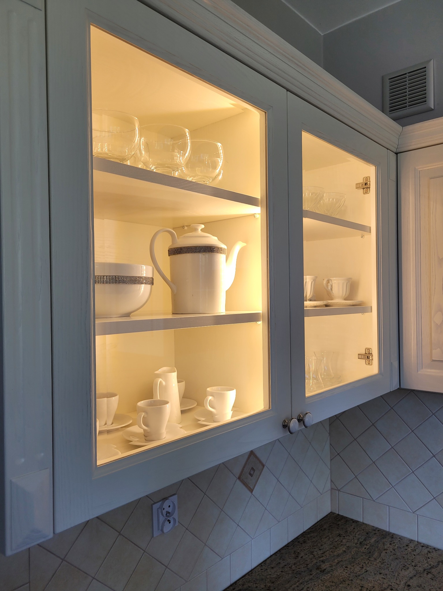 Oświetlona witryna kuchenna z białymi, zdobionymi naczyniami, szklankami i filiżankami na półkach, widoczna płytka ceramiczna na ścianie i gniazdko elektryczne.