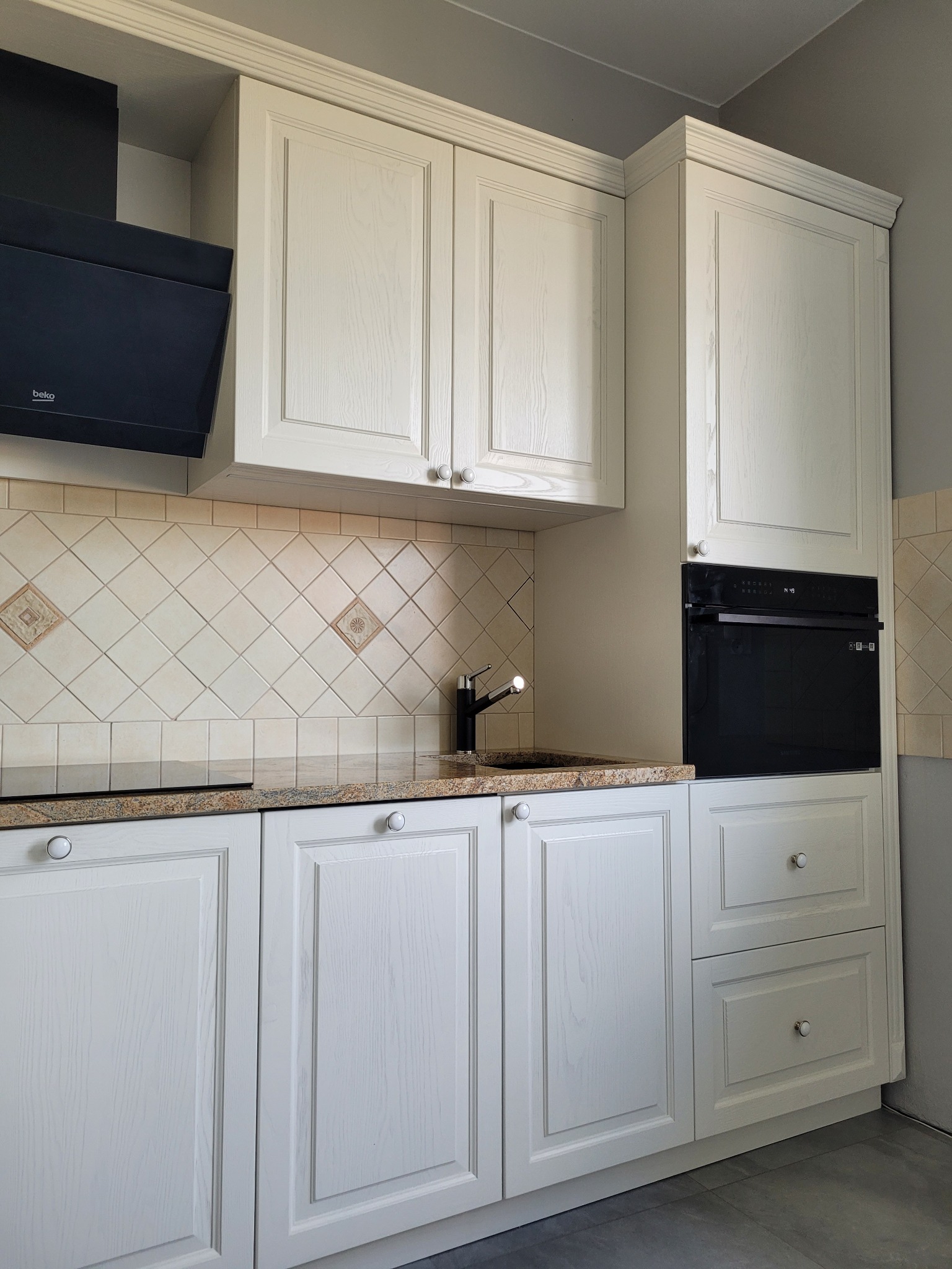 Jasna zabudowa kuchenna z frezowanymi frontami, beżowy blat z delikatnym wzorem i kremowe kafelki z dekoracyjnymi wstawkami, nowoczesny czarny okap i piekarnik.