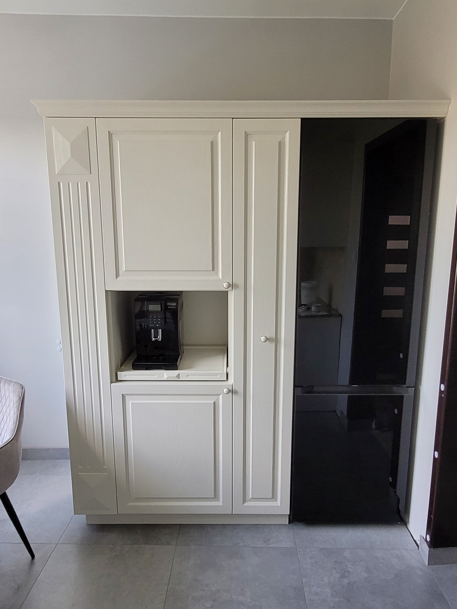 Biała zabudowa kuchenna z wbudowanym ekspresem do kawy i lodówką z czarnym frontem, widok na fragment krzesła.