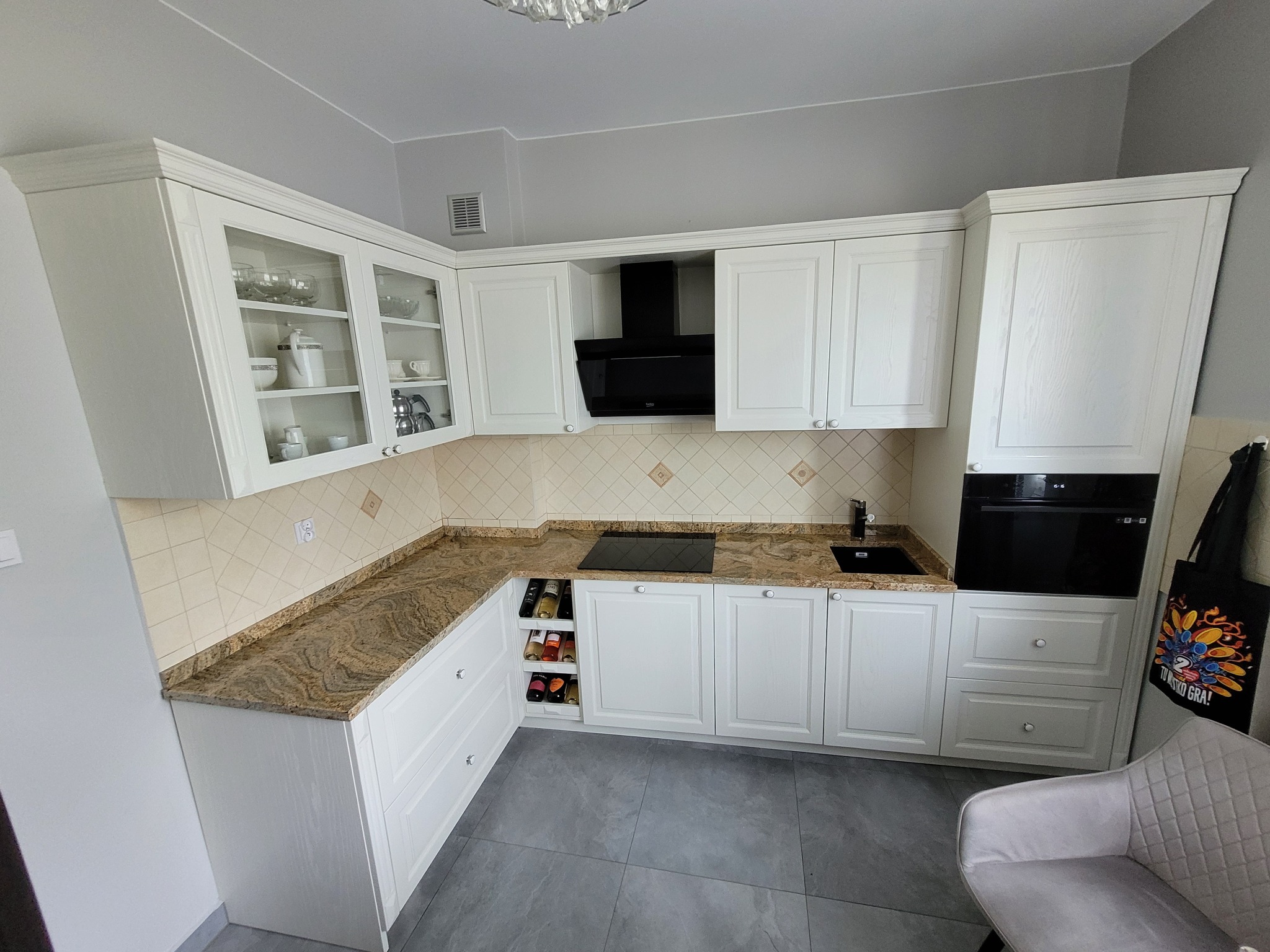 Jasna kuchnia na wymiar z blatem o wzorze marmuru, czarnym okapem i wbudowanym piekarnikiem, widoczne szklane witryny górnych szafek i półka na wino.