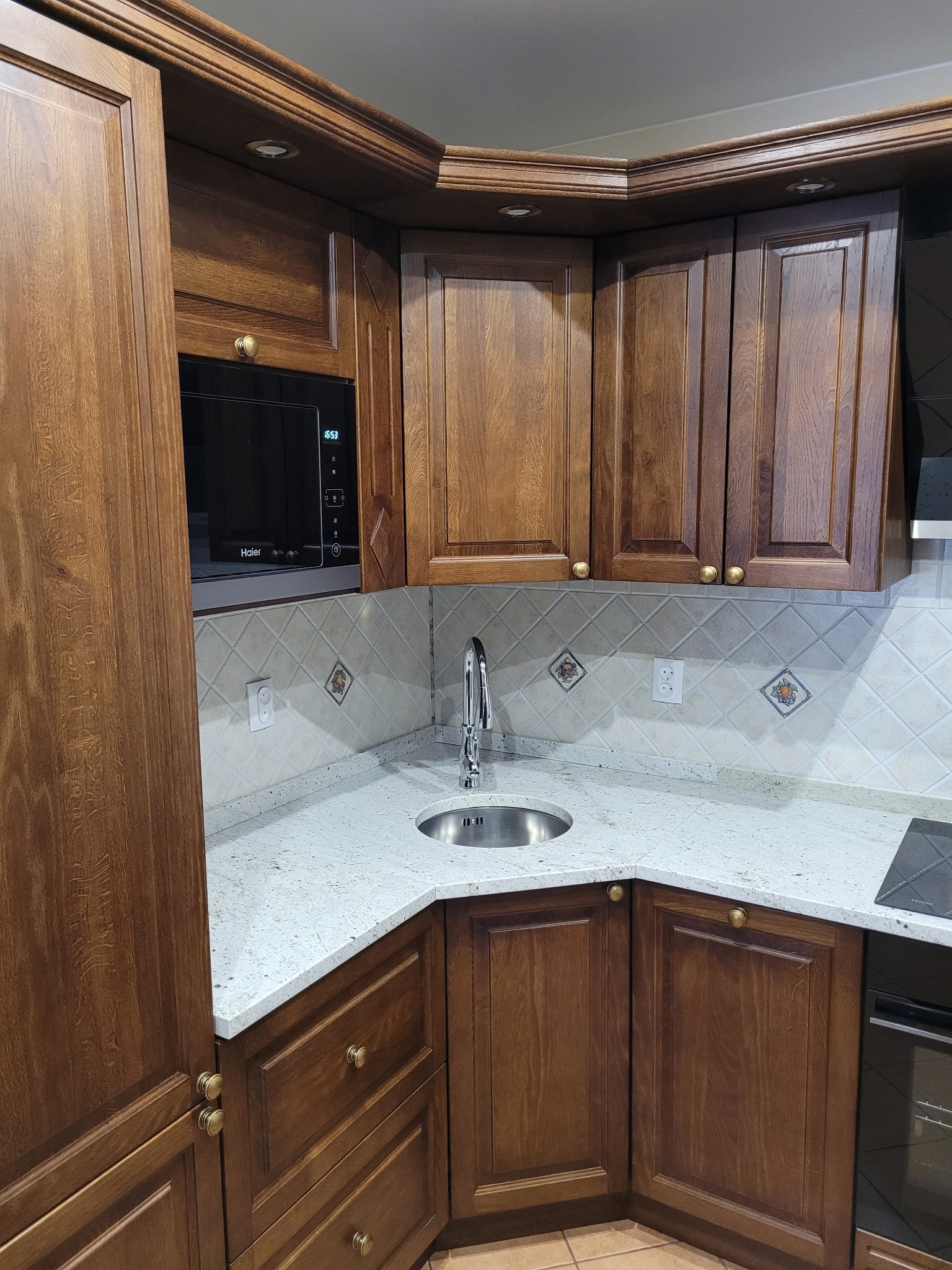 Kąt kuchni z drewnianymi szafkami, blatem z jasnego granitu, okrągłym zlewem ze stali nierdzewnej i mikrofalówką Haier w zabudowie.