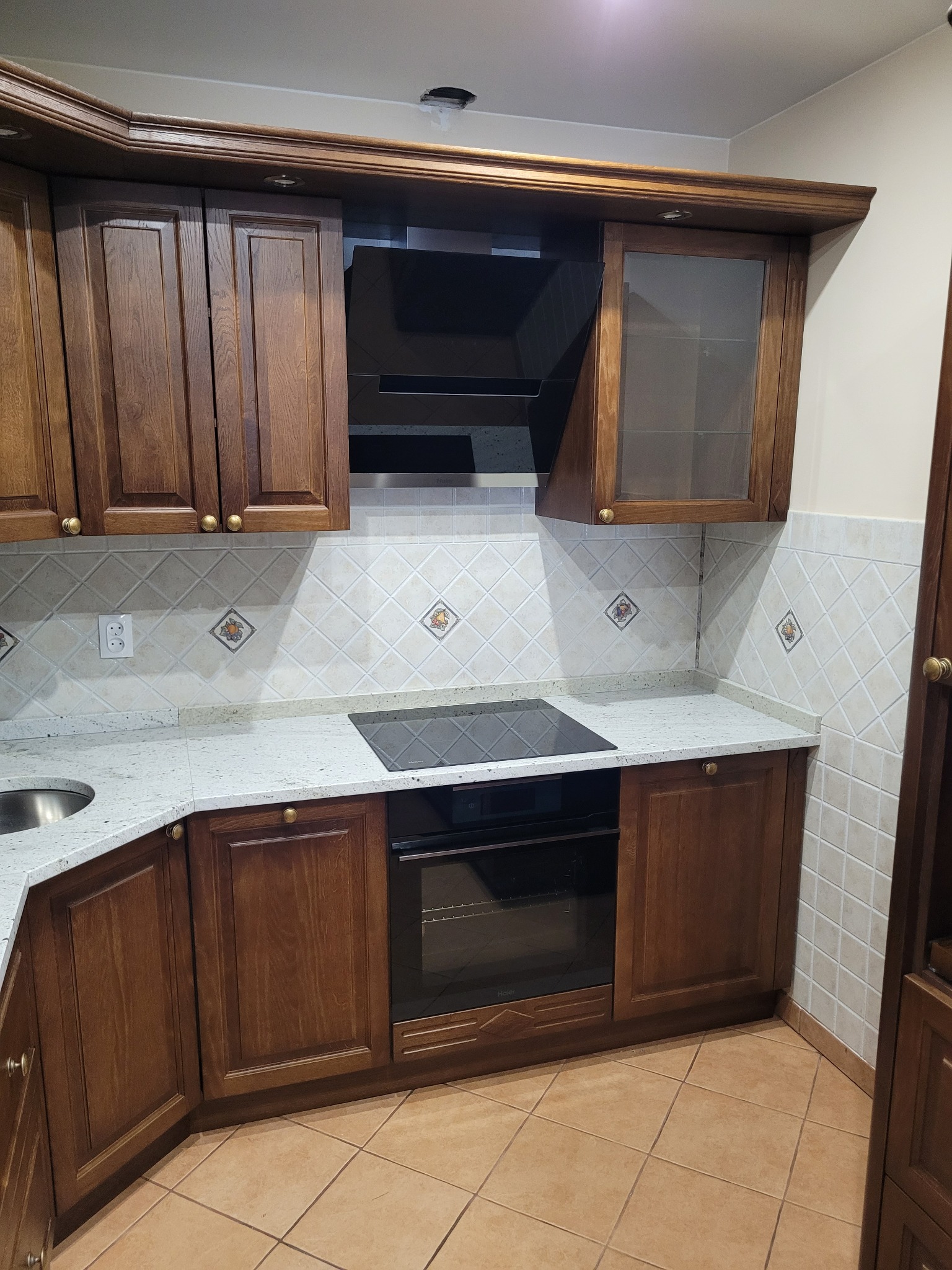 Klasyczna kuchnia z drewnianymi szafkami w odcieniach brązu, jasnym blatem z delikatnym wzorem i płytkami na ścianie z subtelnymi, dekoracyjnymi motywami owoców. Widoczna płyta indukcyjna...