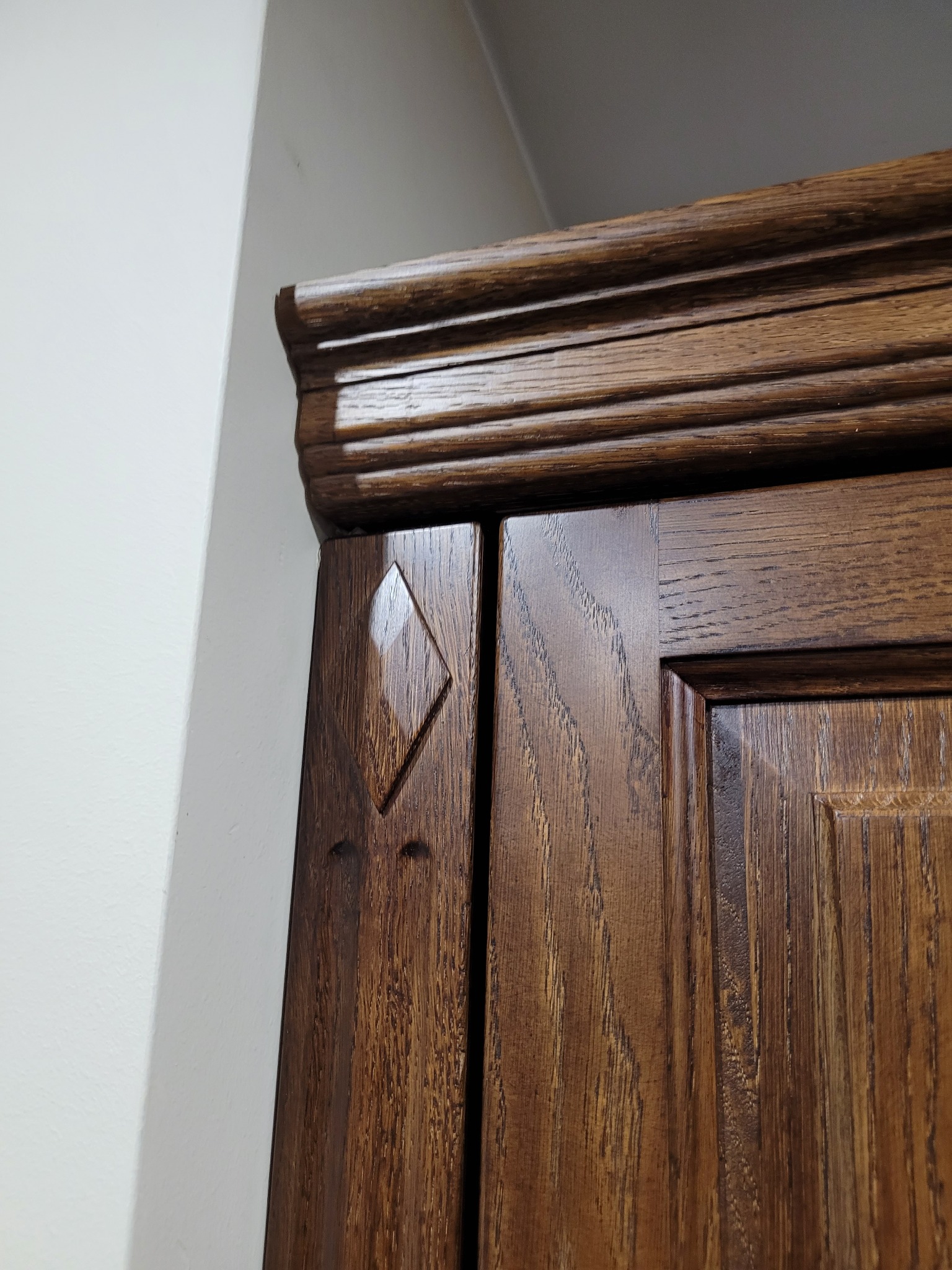 Fragment górnej części ciemnobrązowej drewnianej szafy z dekoracyjnym frezem i romboidalnym elementem na drzwiach, przylegającej do białej ściany.