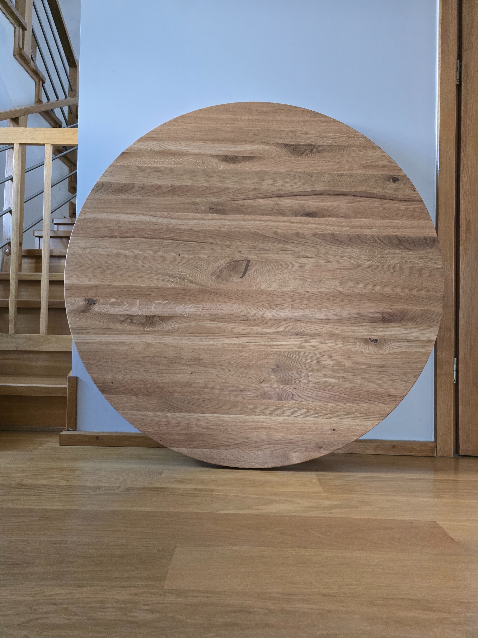 Okrągły, surowy blat z jasnego drewna dębowego, oparty o ścianę, widoczne schody w tle.
