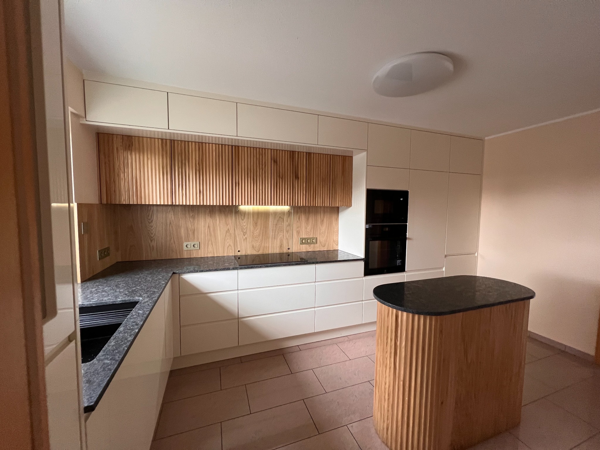 Nowoczesna kuchnia z jasnymi frontami, drewnianymi akcentami na szafkach wiszących i wyspie, czarnym blatem i płytą indukcyjną, oświetleniem podszafkowym i płytkami podłogowymi w odcieniach beżu.