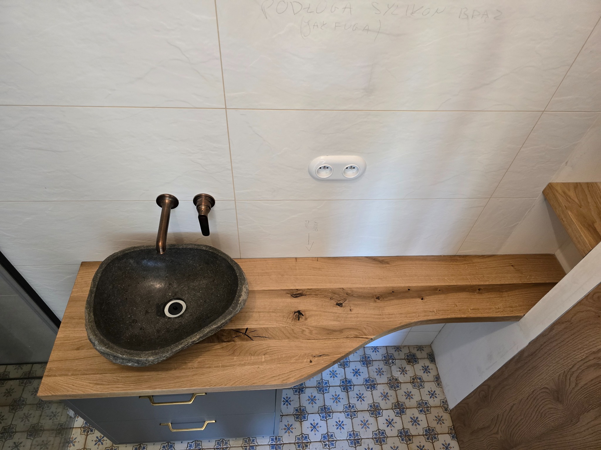 Łazienka z blatem drewnianym i kamienną umywalką. Widoczne białe płytki na ścianie z notatkami i gniazdko elektryczne. Ujęcie z góry, prezentujące unikalny design.