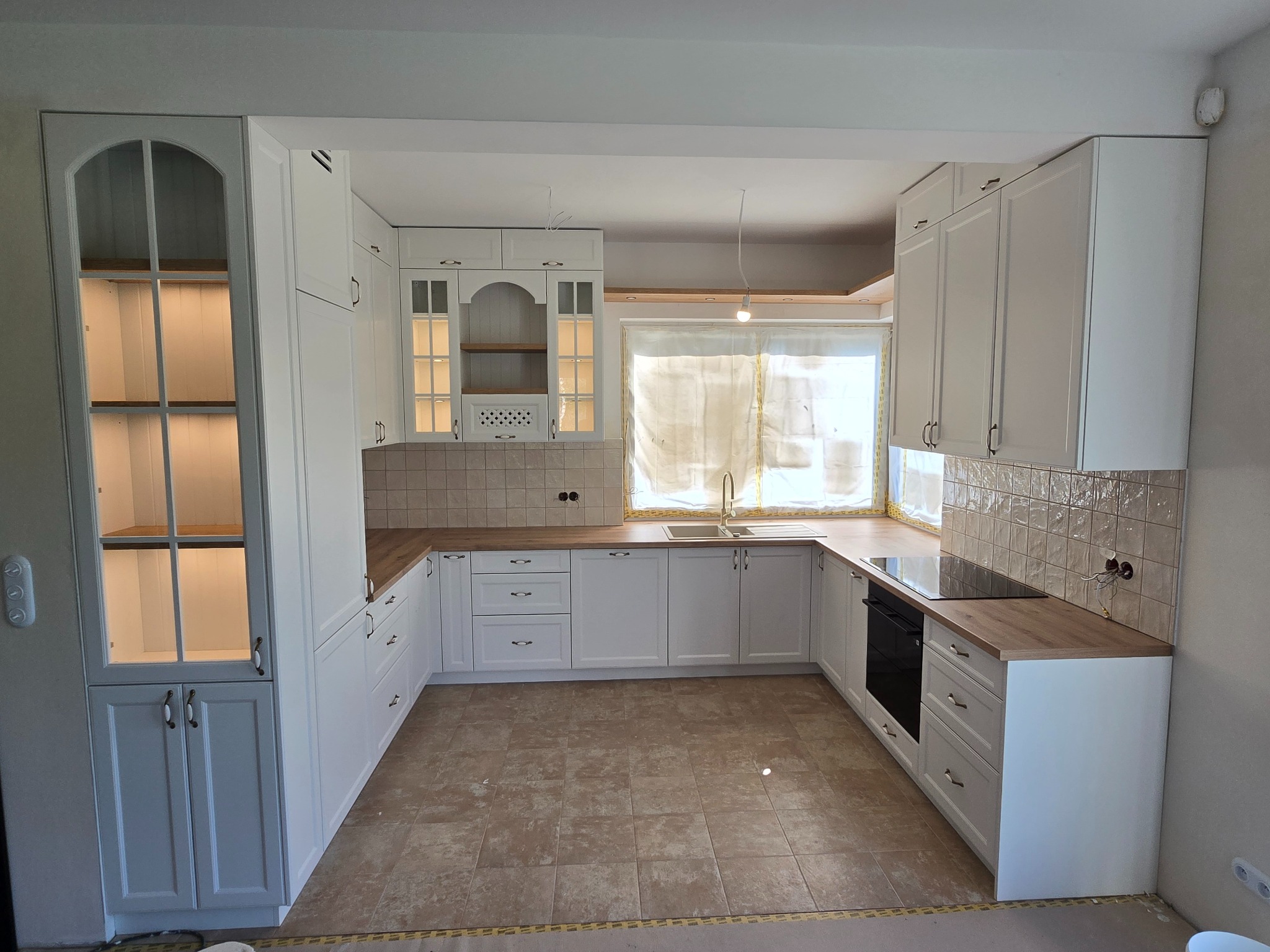 Jasna kuchnia w stylu klasycznym z drewnianymi blatami i przeszklonymi szafkami, oświetlona od wewnątrz, z widocznymi elementami wykończeniowymi.