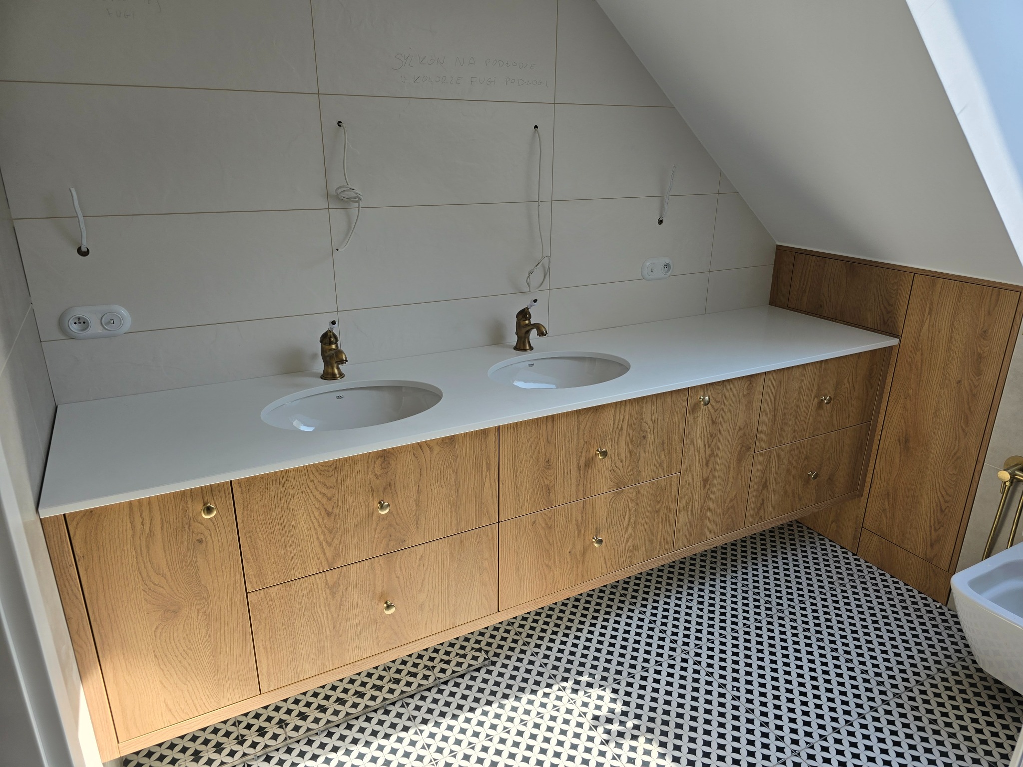 Łazienka na poddaszu z drewnianą szafką pod umywalki i białym blatem, złote krany, wzorzysta podłoga. Widoczne przygotowania do montażu oświetlenia.