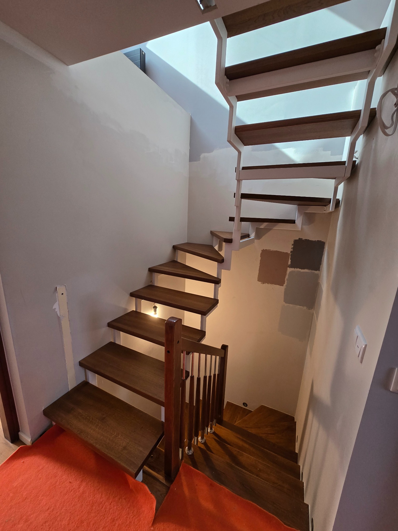 Nowoczesne schody drewniane z białymi elementami konstrukcyjnymi, oświetleniem punktowym na ścianie i próbkami kolorów farb na ścianie, widziane z dołu.