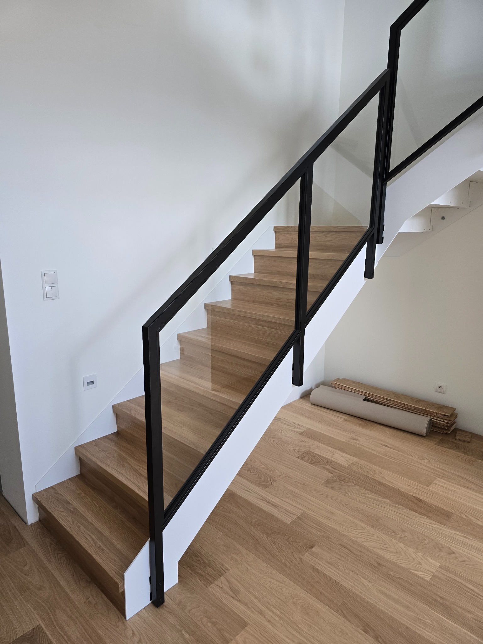 Drewniane schody z czarną balustradą i szklanym wypełnieniem. Na podłodze zwój i deski podłogowe. Widok z dołu na nowoczesne schody w jasnym wnętrzu.