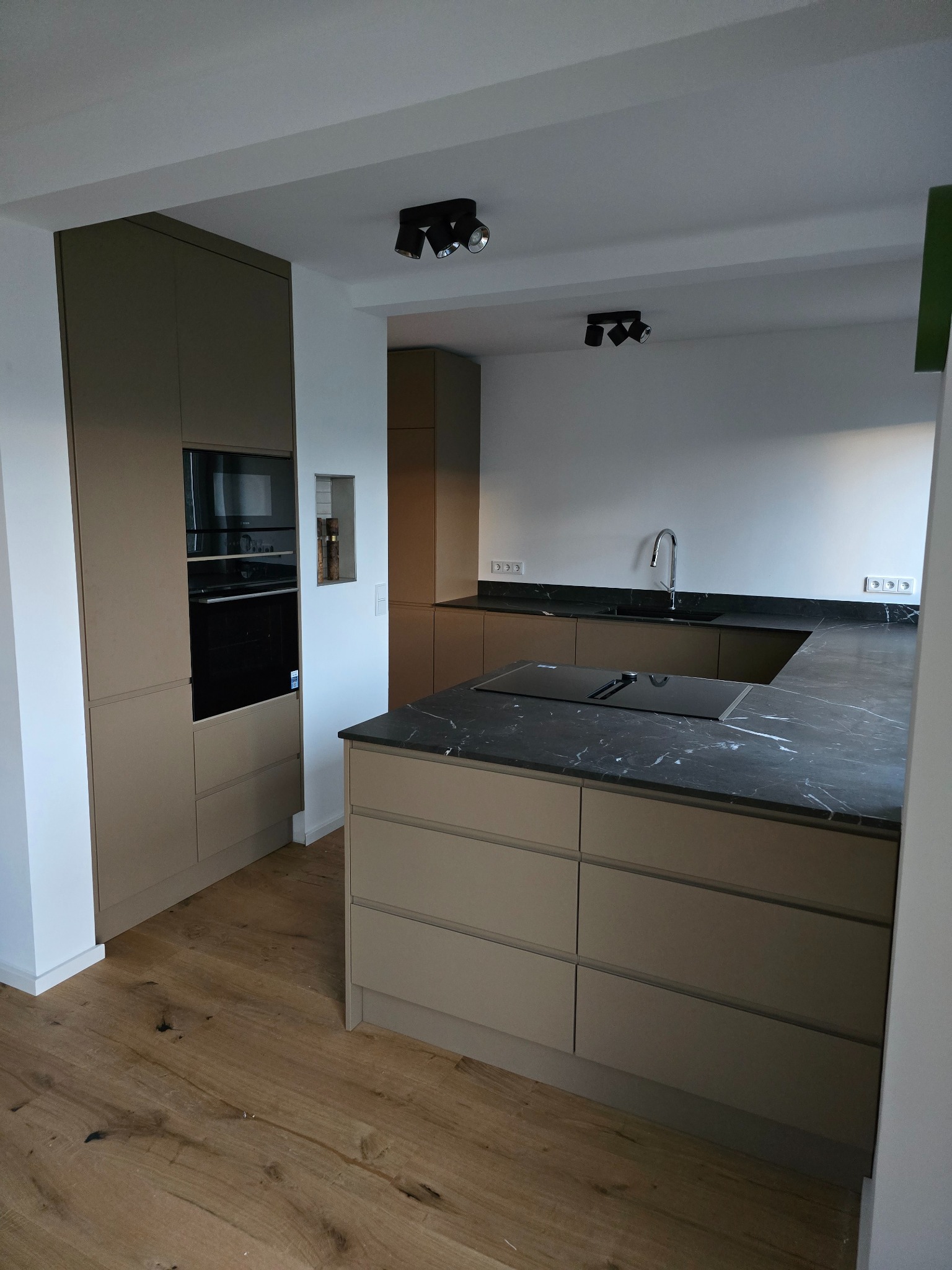 Nowoczesna kuchnia z beżowymi frontami i czarnym blatem z żyłkowaniem. W tle zabudowa z piekarnikiem i płyta indukcyjna na wyspie. Drewniana podłoga i oświetlenie punktowe.