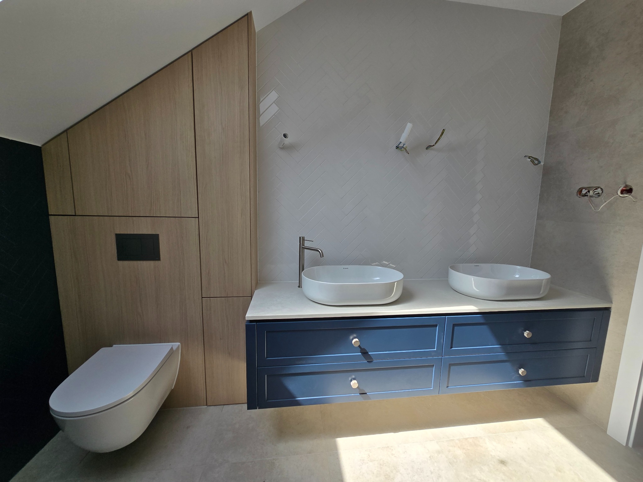 Nowoczesna łazienka z szafką pod umywalki w kolorze granatowym i drewnianą zabudową pod skosem. Umywalki owalne, białe. Na ścianie płytki ułożone w jodełkę.