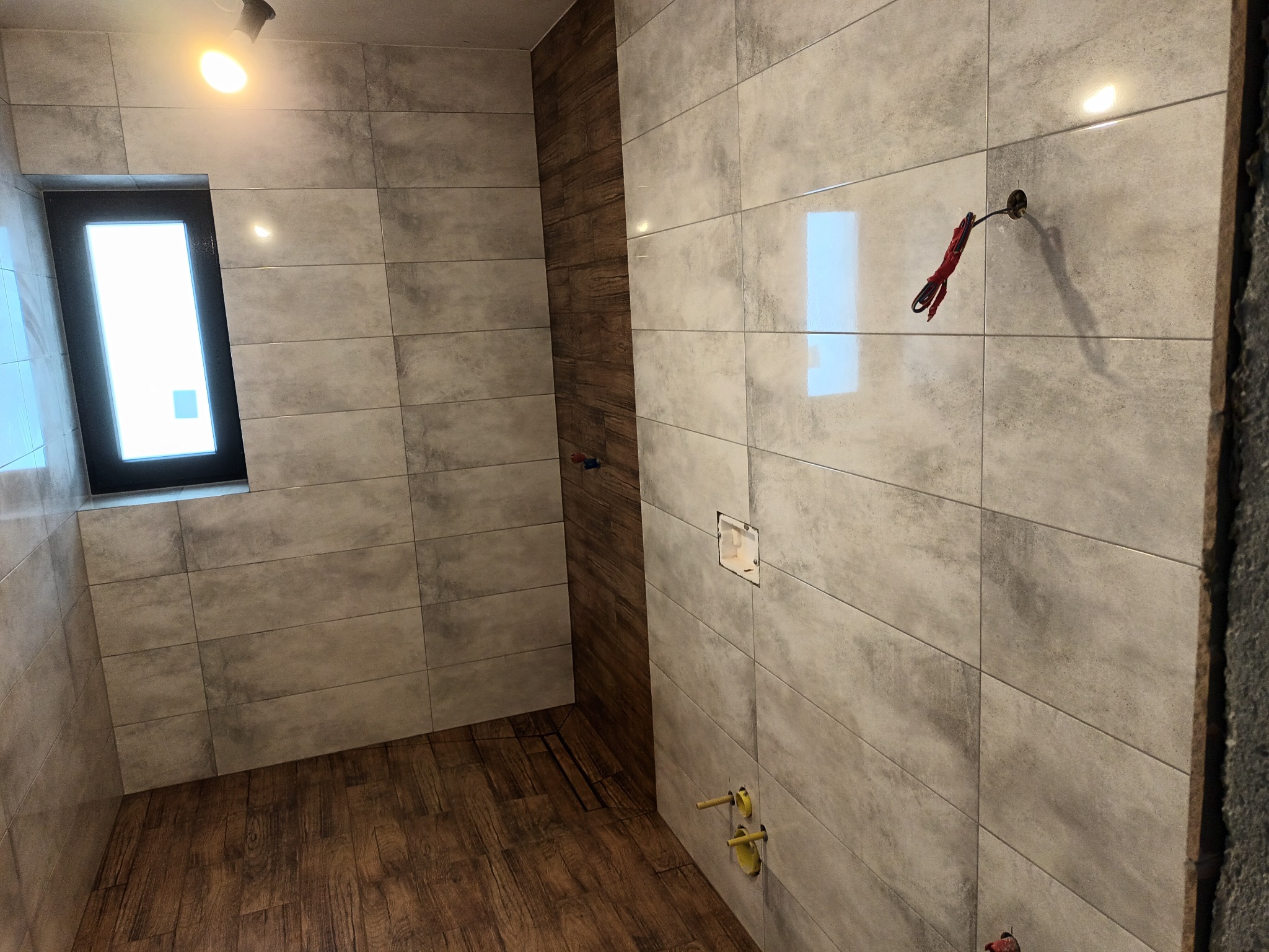 Wnętrze łazienki w trakcie remontu: ułożone szare, podłużne płytki ścienne, fragment ściany z drewnopodobnymi panelami, drewnopodobna podłoga, okno, wystające kable i rury instalacji wodnej.