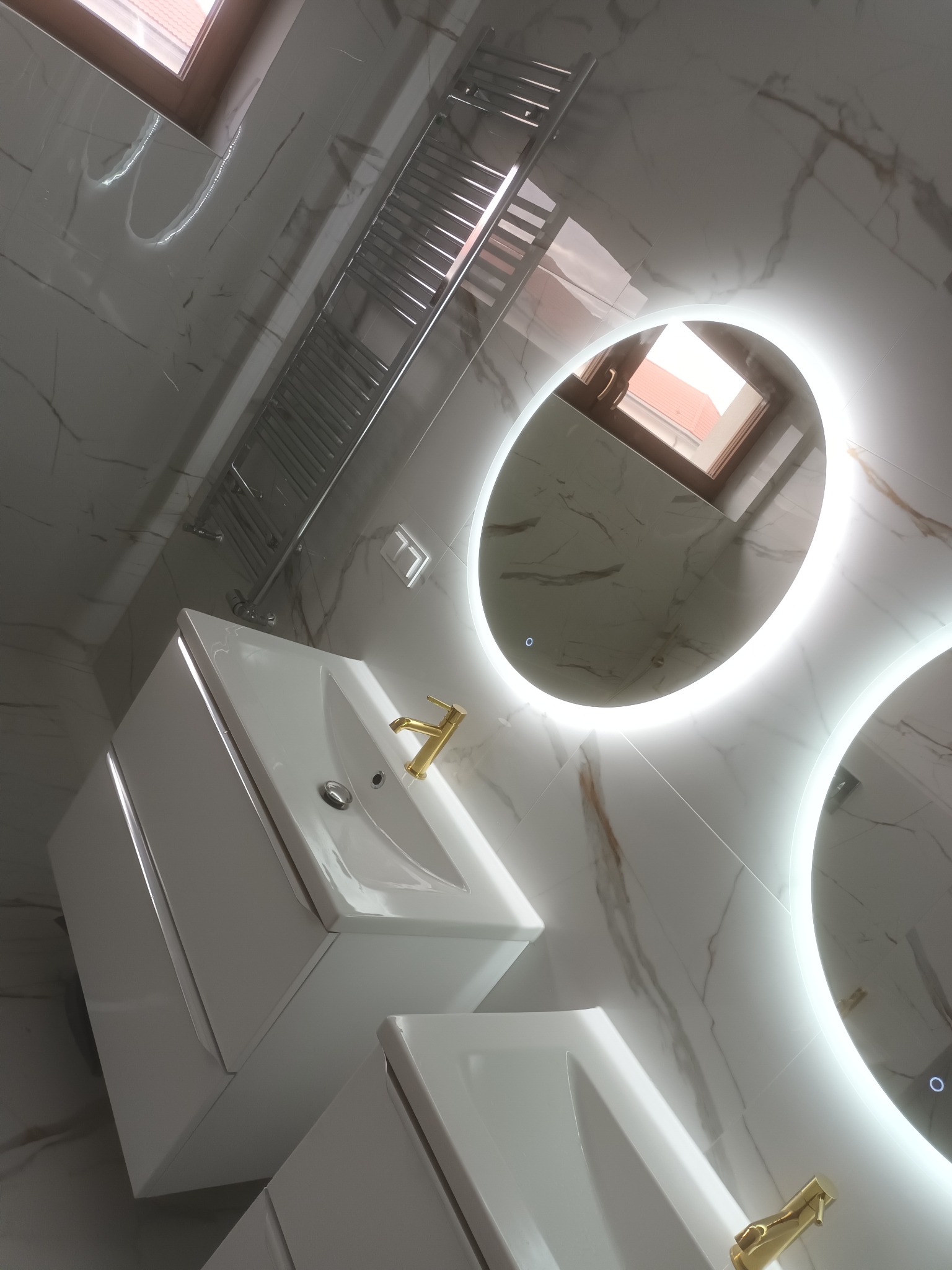 Nowoczesna łazienka z marmurowymi płytkami, podwójną umywalką z szafkami, złotą armaturą i okrągłymi lustrami z podświetleniem LED.