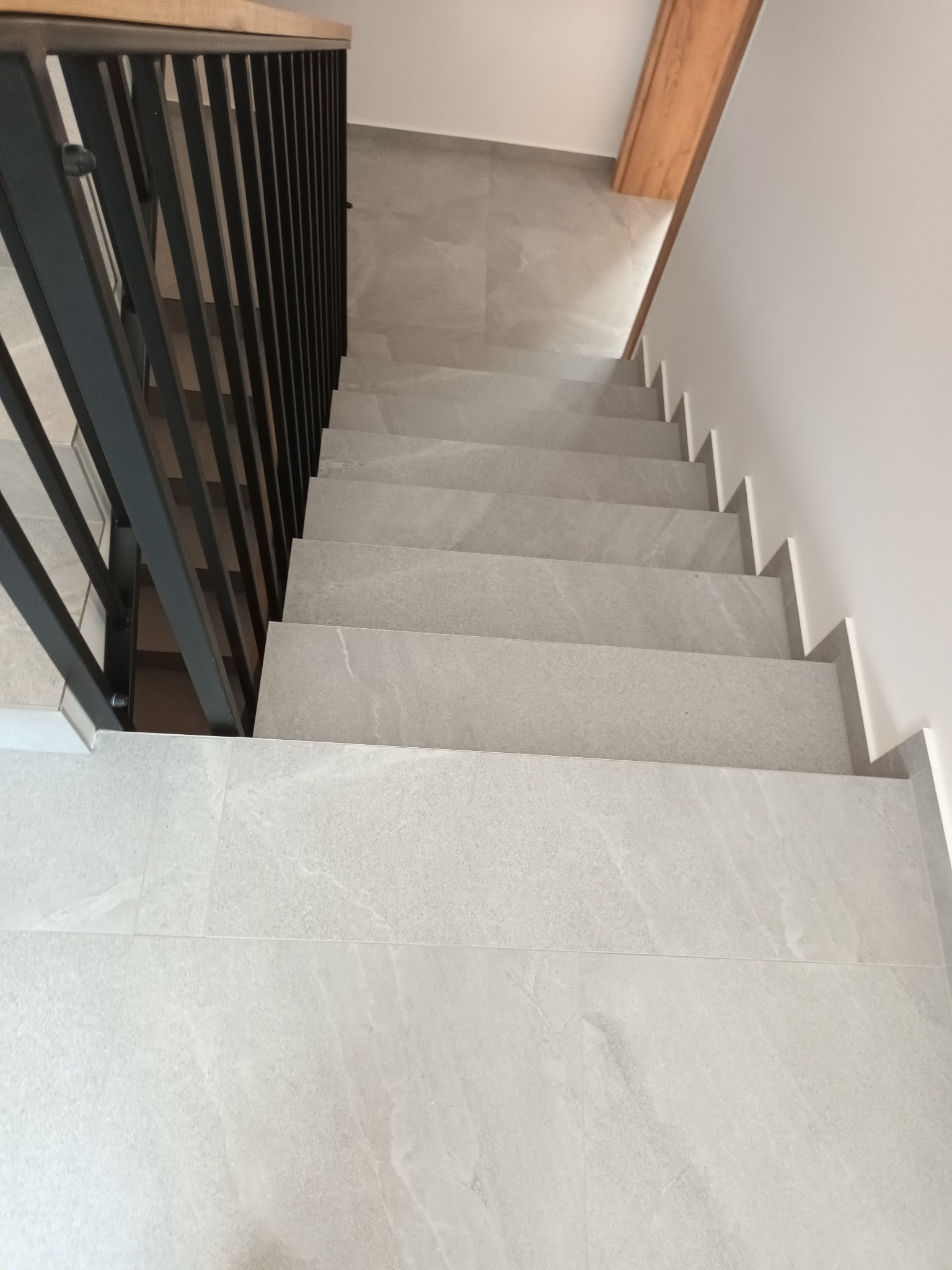 Nowoczesne schody wyłożone jasnoszarą płytką imitującą kamień, z czarną metalową balustradą i drewnianym elementem dekoracyjnym na ścianie.