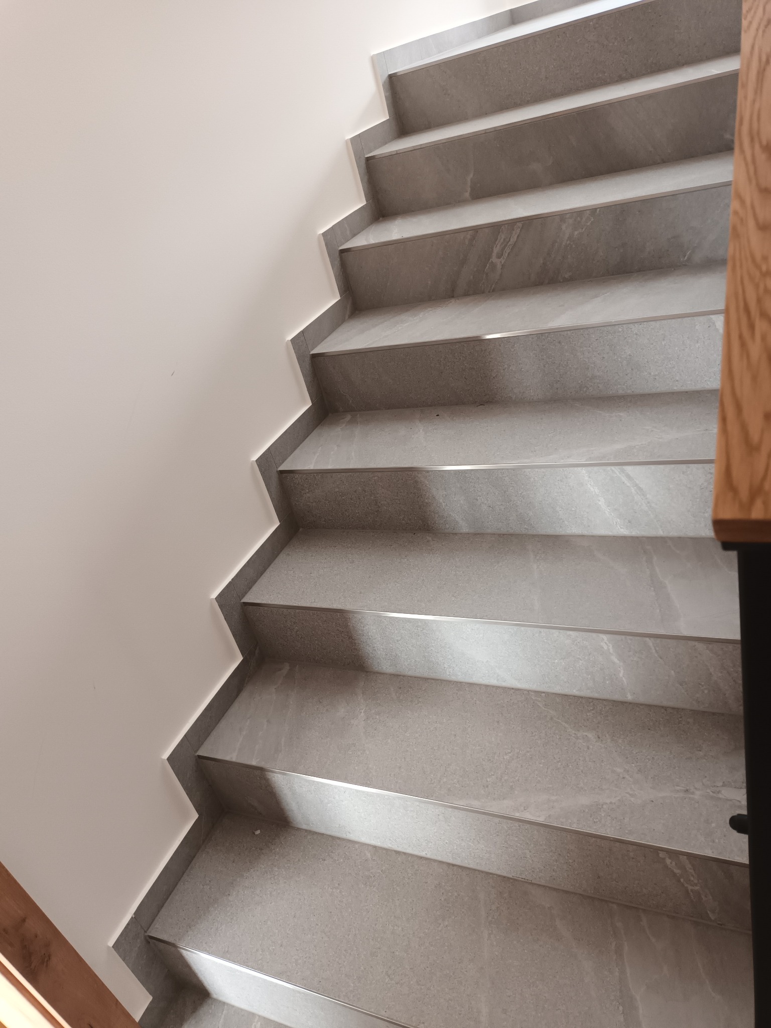 Schody wykończone szarą płytką imitującą kamień, z metalowymi listwami na krawędziach stopni, widoczne z góry w jasnym wnętrzu.