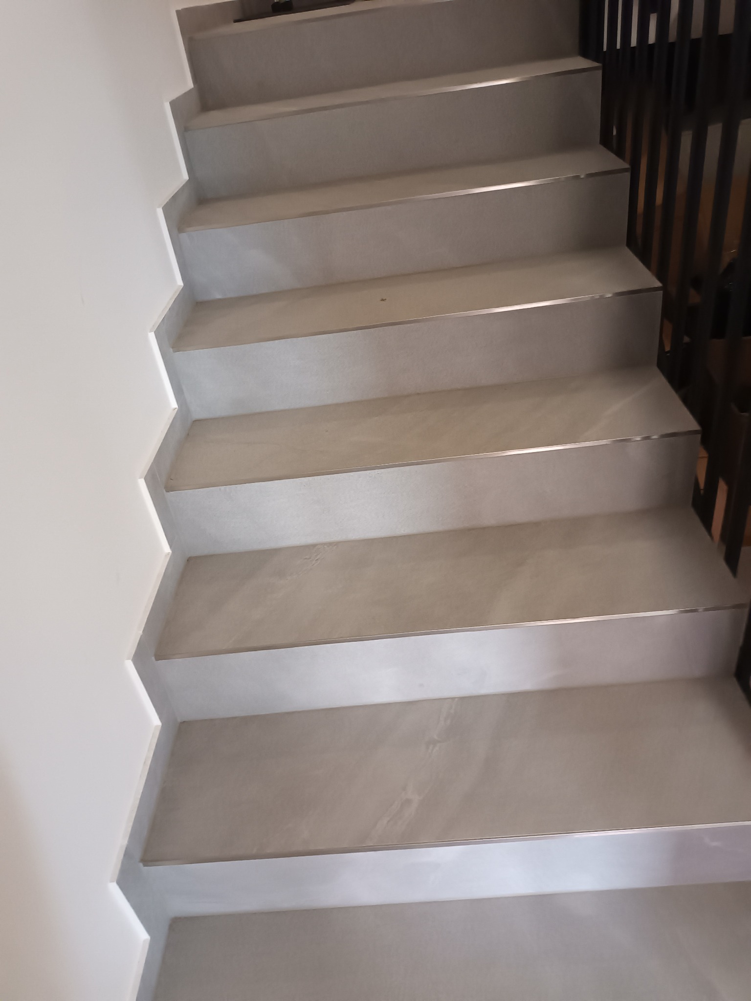 Schody wyłożone szarymi płytkami z metalowymi listwami na krawędziach, widok z dołu na górę, biała ściana po lewej, czarna balustrada po prawej.