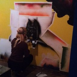 Firma remontowo-wykończeniowa AIRUS sp. z o.o. - Kobieta w kucyku maluje sprayem postać Batmana na żółtej ścianie z motywem klocków Lego, widoczne puszki ze sprayem i telefon.