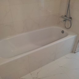 Firma remontowo-wykończeniowa AIRUS sp. z o.o. - Biała wanna obudowana płytkami imitującymi marmur, z zamontowaną baterią prysznicową; widoczne świeżo położone płytki podłogowe z marmurowym wzorem oraz fragment surowej ściany po remoncie.