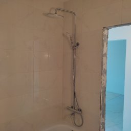 Firma remontowo-wykończeniowa AIRUS sp. z o.o. - Wykończona łazienka z beżowymi płytkami imitującymi marmur, chromowana bateria prysznicowa z deszczownicą i słuchawką, widoczny fragment wanny i surowy otwór drzwiowy.