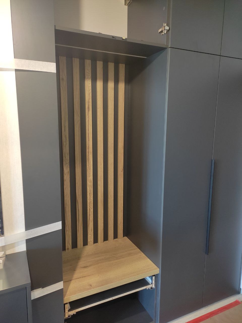 Wnęka z drewnianymi lamelami i siedziskiem z płyty meblowej w zabudowie garderoby w kolorze grafitowym, widoczny fragment listwy przypodłogowej.