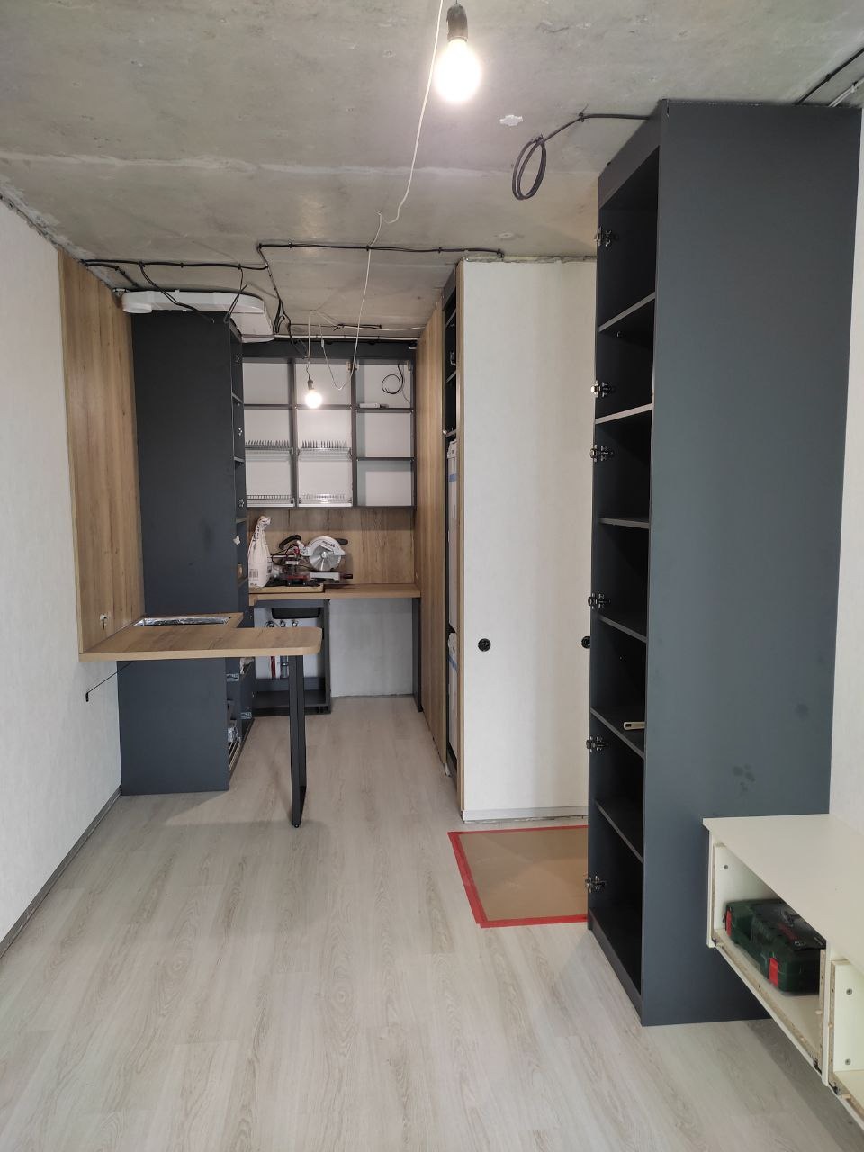 Wnętrze pokoju w trakcie remontu z widocznymi szafami w zabudowie, rozłożonym blatem roboczym, narzędziami i odsłoniętym betonowym sufitem z instalacją elektryczną.