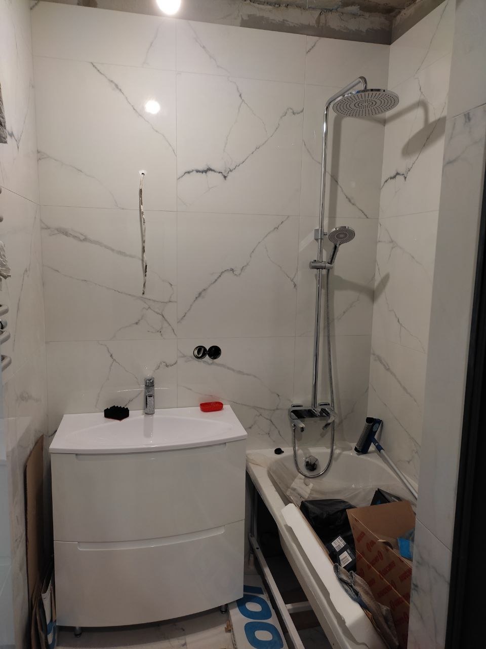 Nowa łazienka w trakcie wykańczania: białe płytki imitujące marmur, biała szafka z umywalką, bateria, zestaw prysznicowy z deszczownicą. Widoczne przygotowania do montażu wanny, kartony i narzędzia...