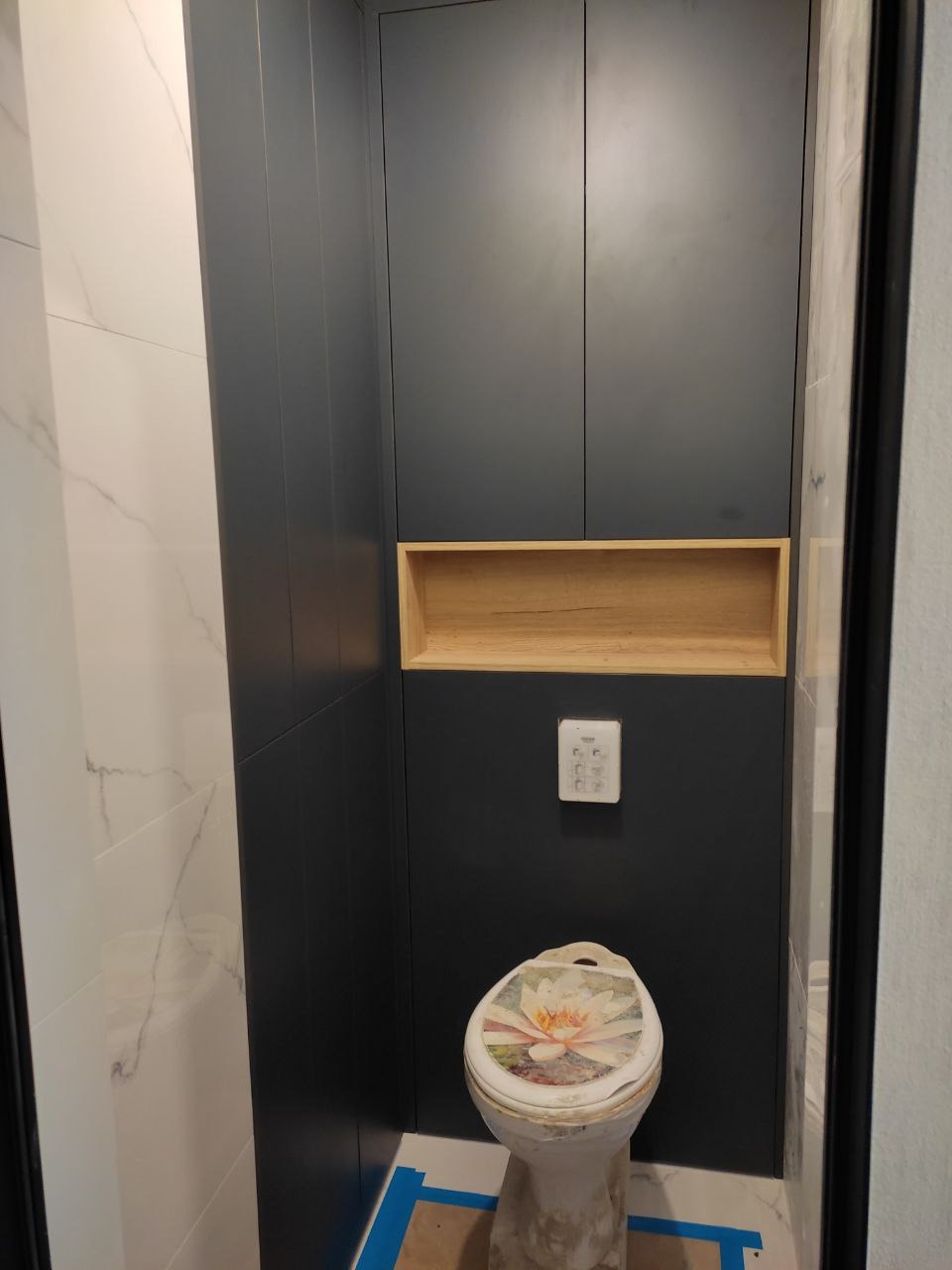 Wąska toaleta po renowacji z zabudową szafkową w kolorze grafitowym, drewnianą wnęką nad spłuczką i płytkami imitującymi marmur na ścianach. Na desce sedesowej naklejka z motywem kwiatowym.