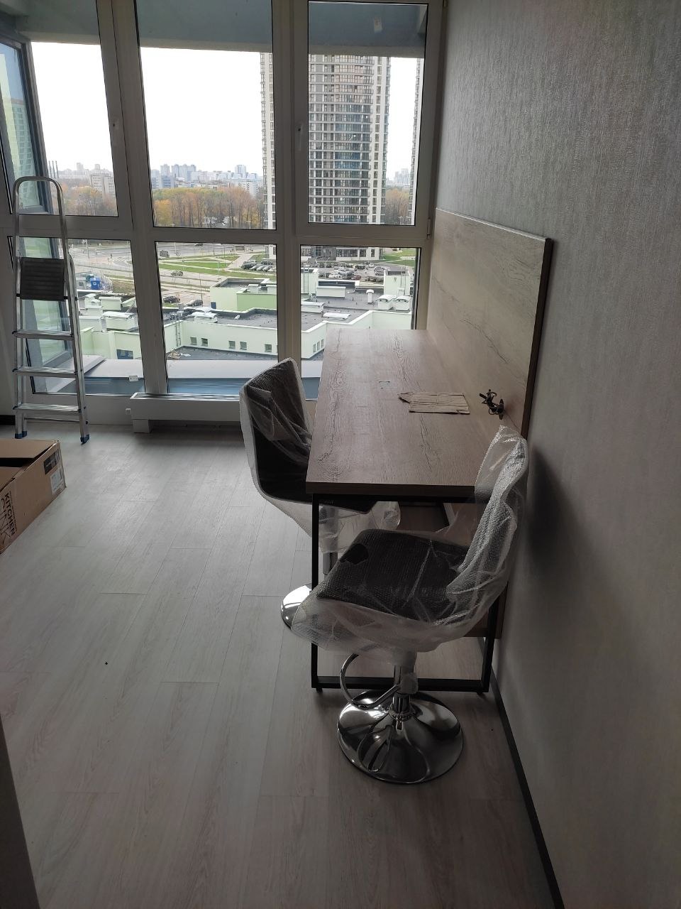 Nowe krzesła barowe i stół zabezpieczone folią ochronną w świeżo wykończonym wnętrzu z dużym oknem i widokiem na miasto, drabina oparta o ścianę.