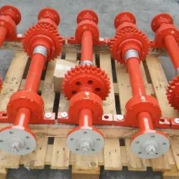 Pomarańczowe, metalowe wały z zębatkami i kołnierzami, ułożone na drewnianej palecie, prawdopodobnie elementy maszyn.