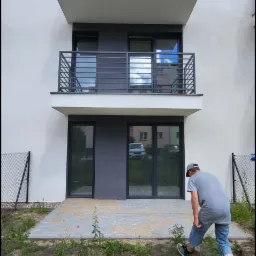 Nowoczesny budynek wielorodzinny z balkonami i wybrukowanym tarasem na parterze, gdzie mężczyzna w dżinsowych szortach pielęgnuje teren zielony.