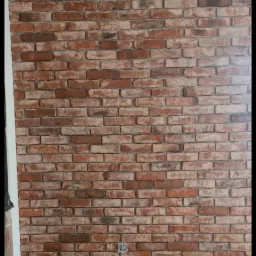 Ściana wykończona panelami imitującymi czerwoną cegłę z widocznym gniazdkiem elektrycznym, na tle surowej, szarej podłogi i białej ściany. Widoczne drobne niedociągnięcia montażu paneli.