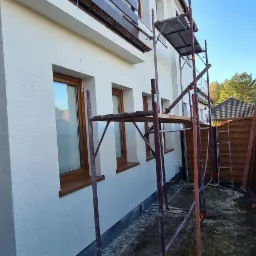 Dom jednorodzinny z nową, jasną elewacją, widoczne brązowe ramy okienne i brązowa balustrada balkonu, ustawione rusztowanie malarskie i rozłożona plandeka na ziemi, ujęcie z dołu.