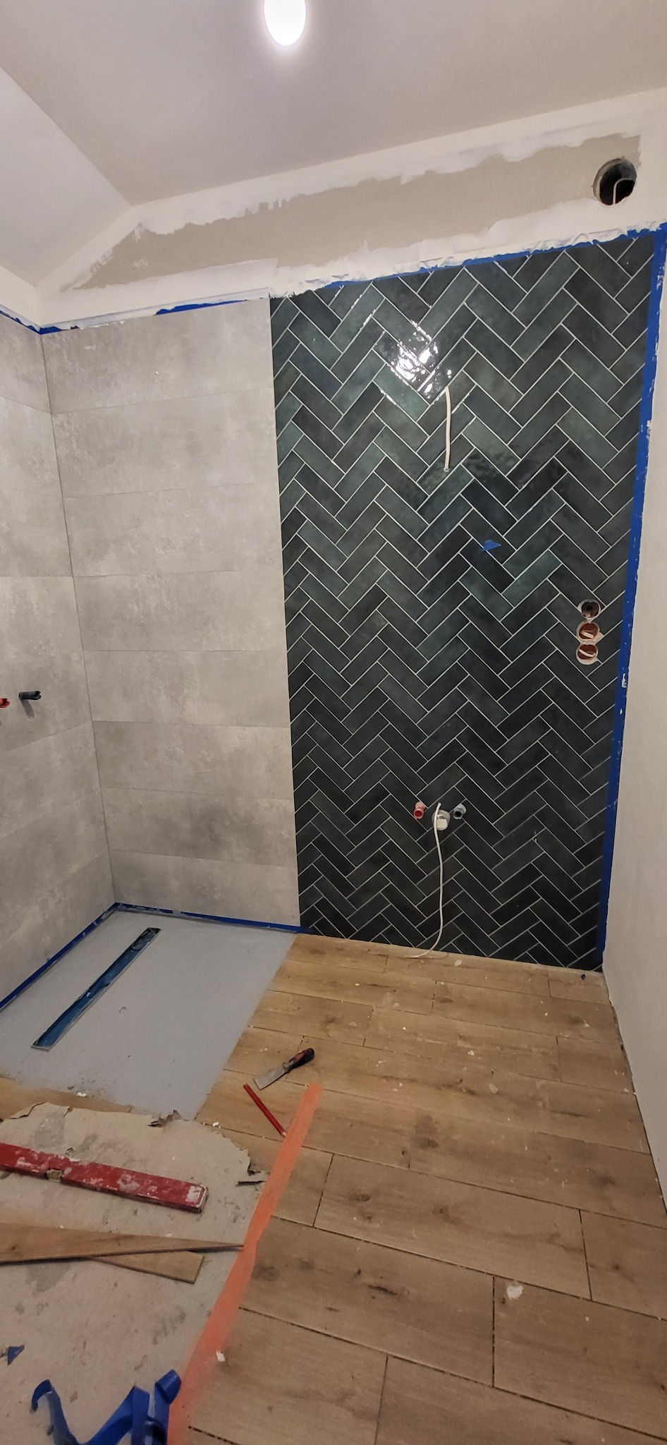 Fragment łazienki w trakcie remontu: ułożone płytki podłogowe imitujące drewno, ściana częściowo wykończona szarymi płytkami, a częściowo ciemnymi płytkami ułożonymi w jodełkę, widoczne narzędzia...