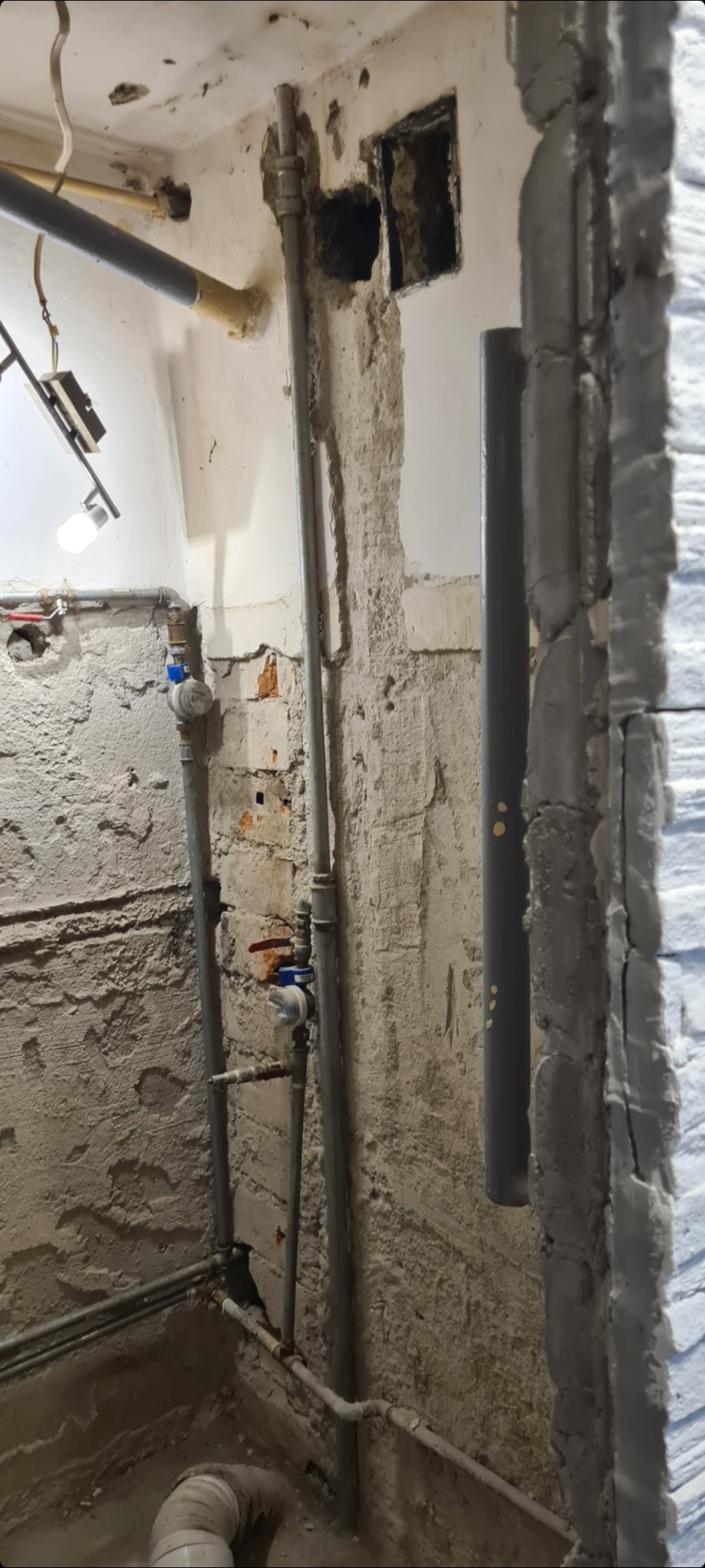 Surowe wnętrze w trakcie remontu z widocznymi rurami, instalacjami i fragmentami otynkowanych ścian z cegły.