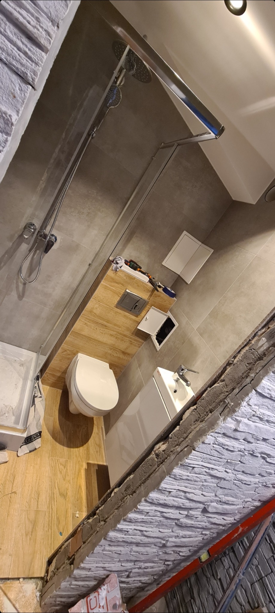 Wnętrze łazienki w trakcie remontu, widoczna kabina prysznicowa, toaleta, umywalka, płytki imitujące drewno na podłodze i ścianie, oraz dekoracyjna ściana z imitacji kamienia w trakcie montażu.