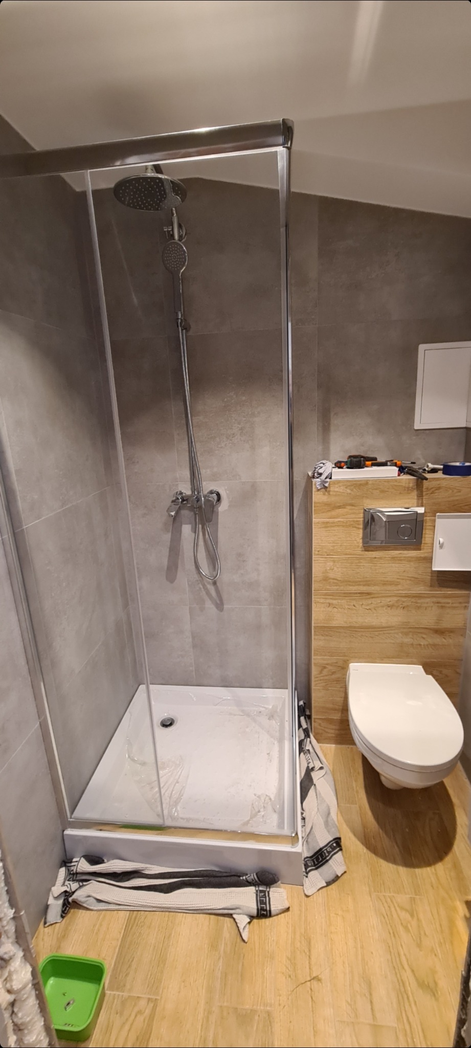 Nowoczesna kabina prysznicowa z szarymi płytkami i drewnopodobną zabudową toalety, widoczne narzędzia i ręczniki sugerują prace wykończeniowe w łazience.