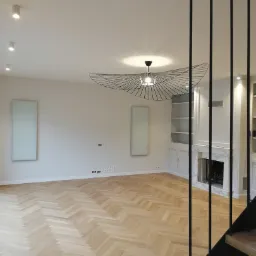 Jasne, przestronne wnętrze po remoncie z parkietem ułożonym w jodełkę, ozdobnym żyrandolem i minimalistycznym kominkiem z marmurowym obramowaniem, widok z perspektywy schodów.