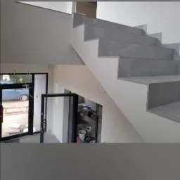 Wykończone schody wewnętrzne z szarymi płytkami, widoczne z góry, z fragmentem otwartego pomieszczenia z wiadrami i narzędziami w tle.