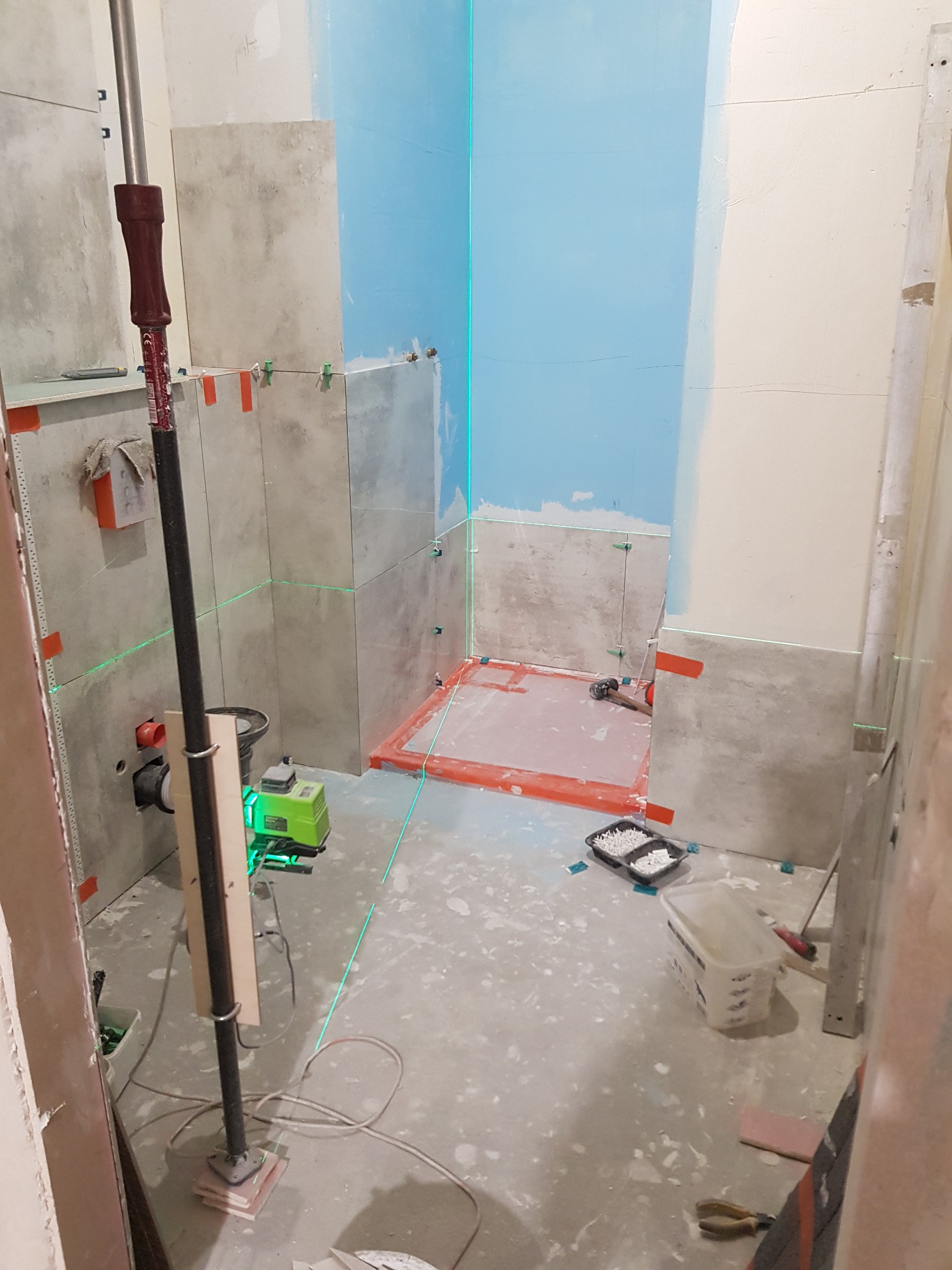 Montaż płytek w łazience z użyciem poziomicy laserowej, widoczne krzyżyki dystansowe i elementy instalacji wodnej