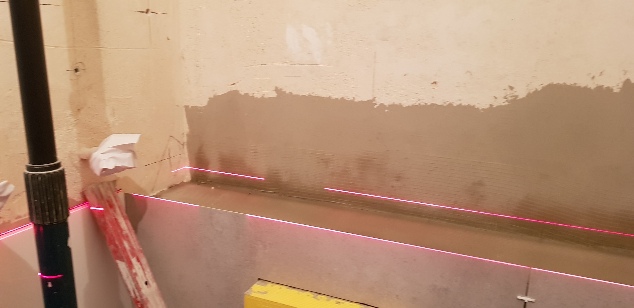 Przygotowanie ściany do położenia płytek z użyciem lasera poziomującego, widoczna warstwa wyrównująca i zaznaczone linie cięcia.