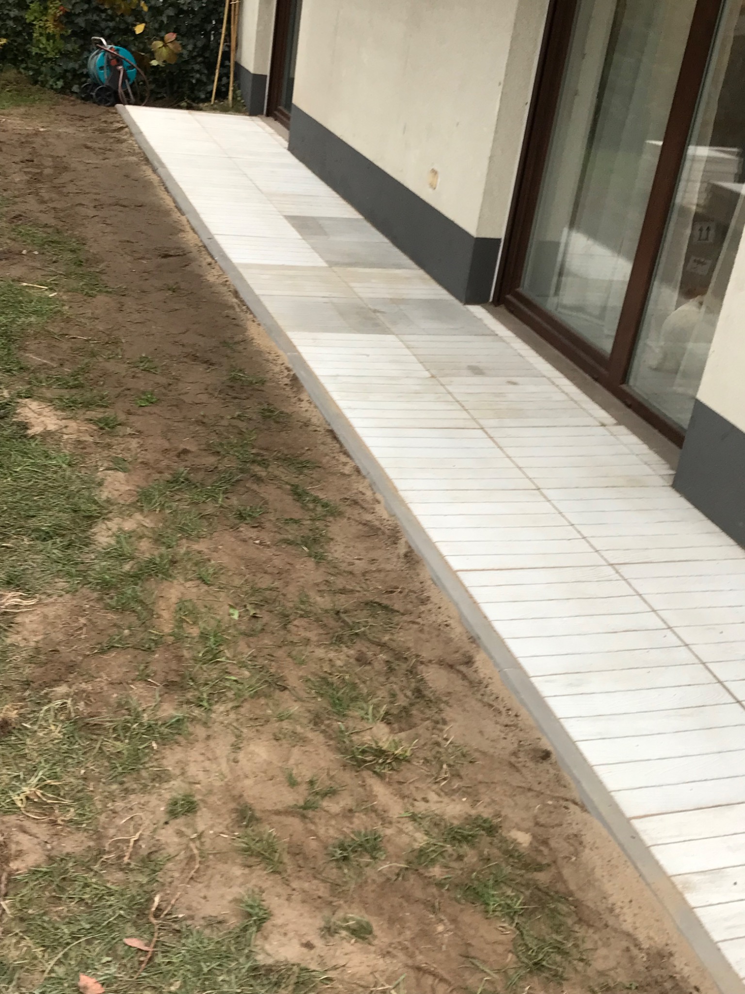 Ułożona, jasna kostka brukowa wzdłuż ściany budynku w Warszawie, widoczny fragment trawnika i narzędzia ogrodnicze w tle.