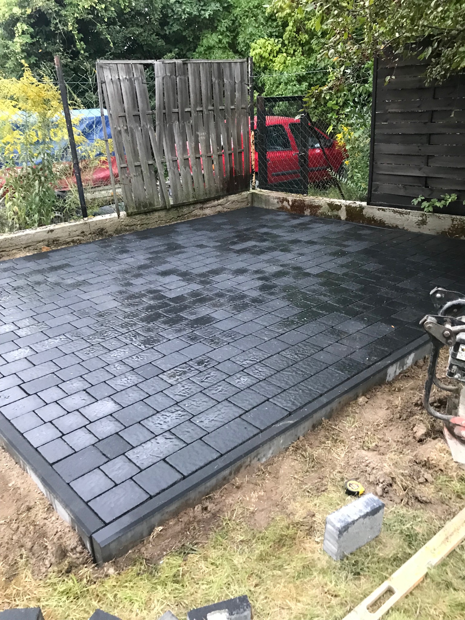Ułożona, mokra, ciemnoszara kostka brukowa na przygotowanym podłożu, widoczna zagęszczarka i miarka, w tle ogrodzenie i zieleń.