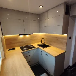 Nowoczesna, jasna kuchnia z drewnianymi blatami i szarymi, błyszczącymi szafkami, oświetleniem LED pod szafkami wiszącymi i czarną armaturą, widoczna płyta grzewcza i gniazdka elektryczne w blacie.