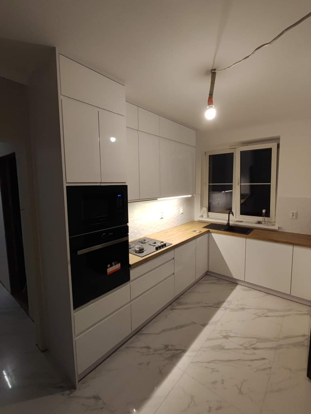 Nowoczesna kuchnia z białymi, lakierowanymi frontami, drewnianym blatem i marmurową podłogą, oświetlona wiszącą żarówką na kablu.